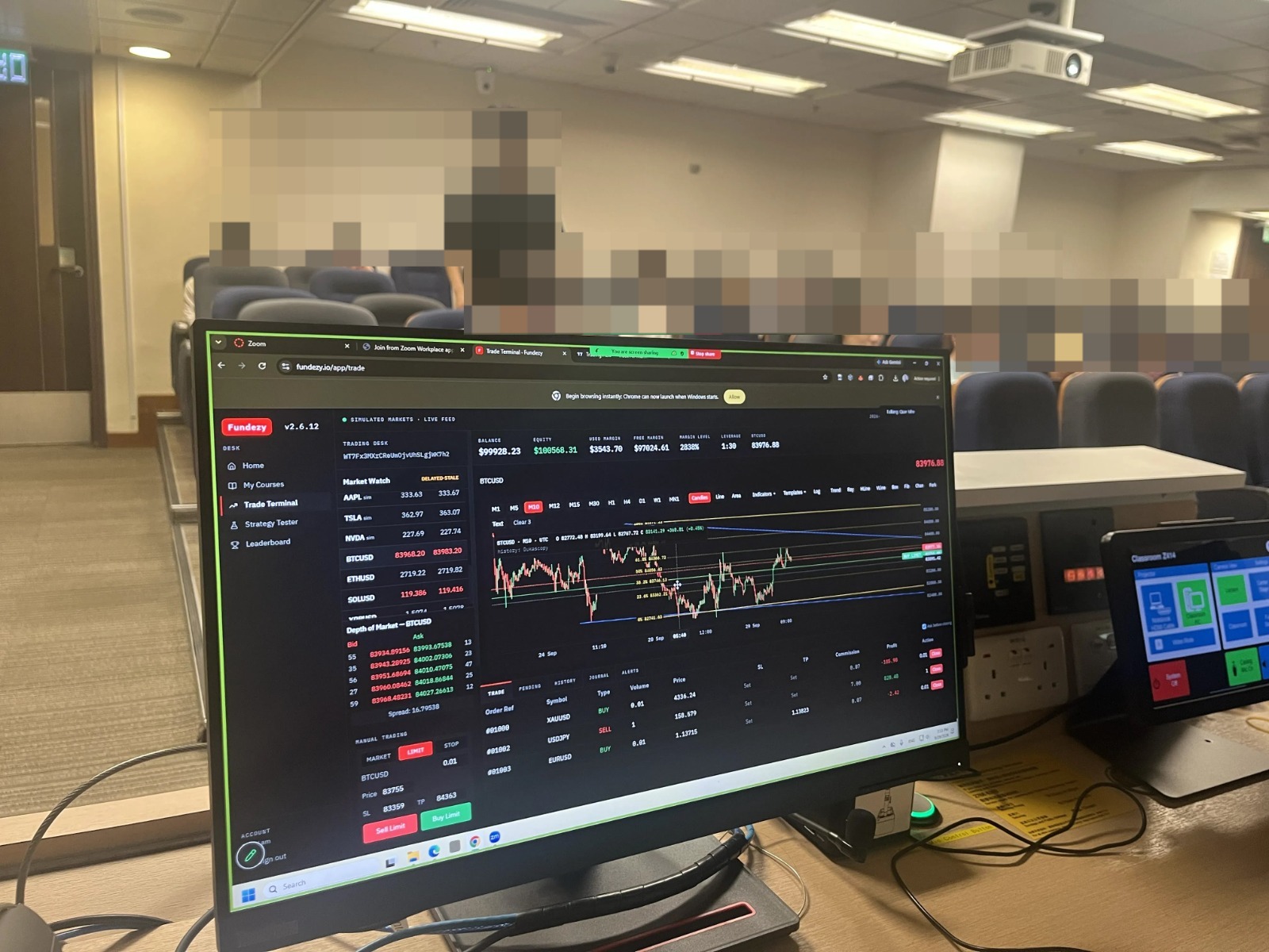 A Fundezy trading desk on the podium monitor at the front of a university classroom in Hong Kong — trade terminal with a watchlist, a BTC/USD candlestick chart and an order ticket, students at their seats in the background.